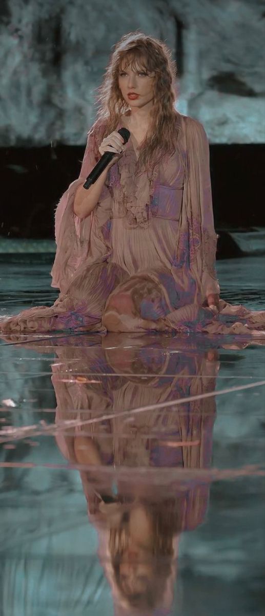 a woman sitting in the water holding a microphone and wearing a pink dress with long sleeves