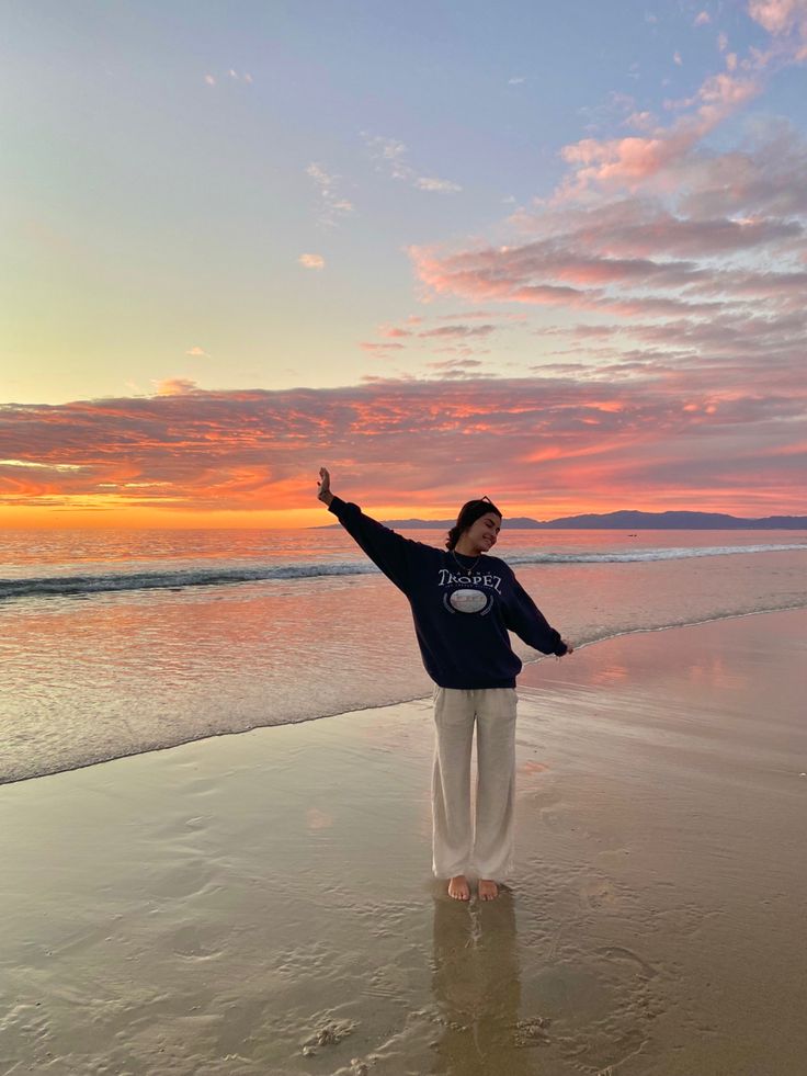 a woman standing on top of a sandy beach next to the ocean at sunset with her arms in the air