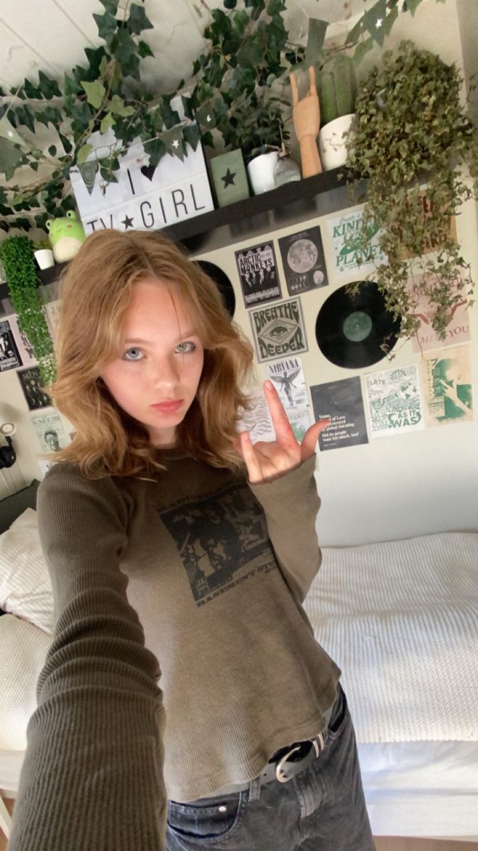 a woman standing in front of a bed holding her hand up to the camera and making a peace sign