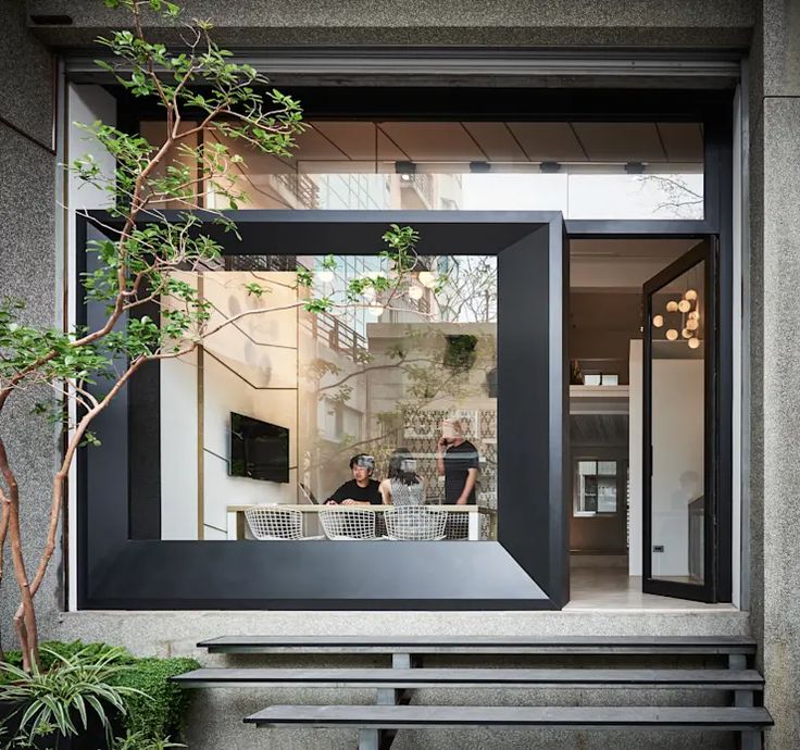 two people sitting at a table in front of a large window with plants on it