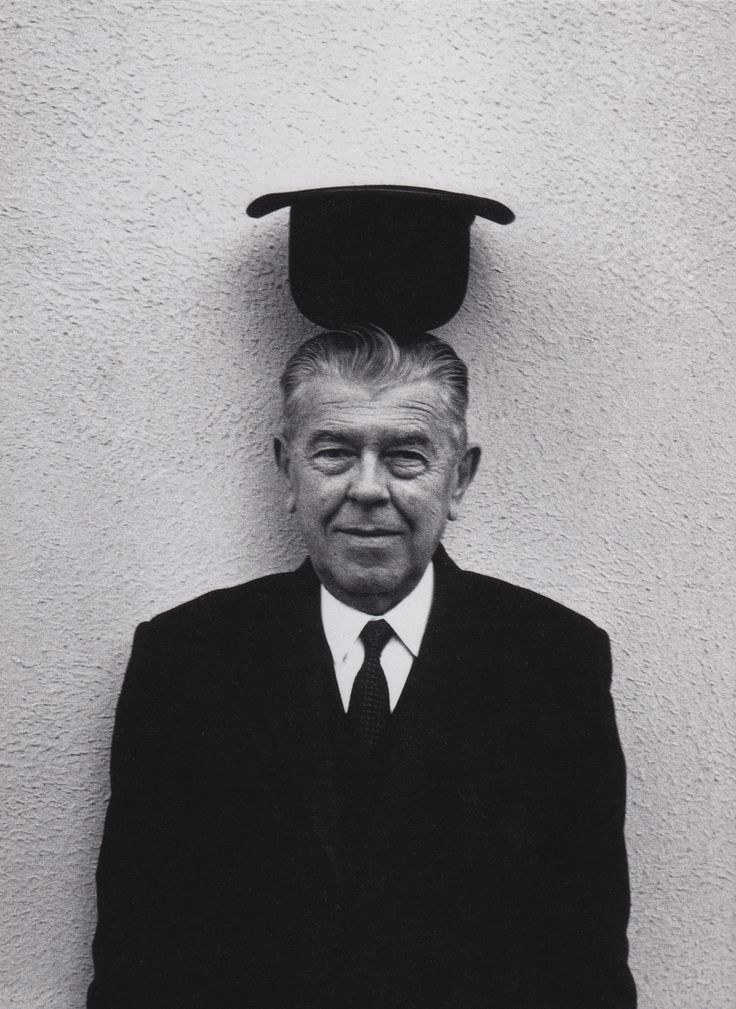 an old man in a suit and tie with a graduation cap on his head, standing against a white wall