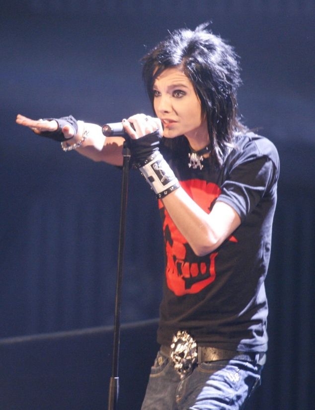 a man with black hair holding a microphone in his right hand and wearing a skull t - shirt
