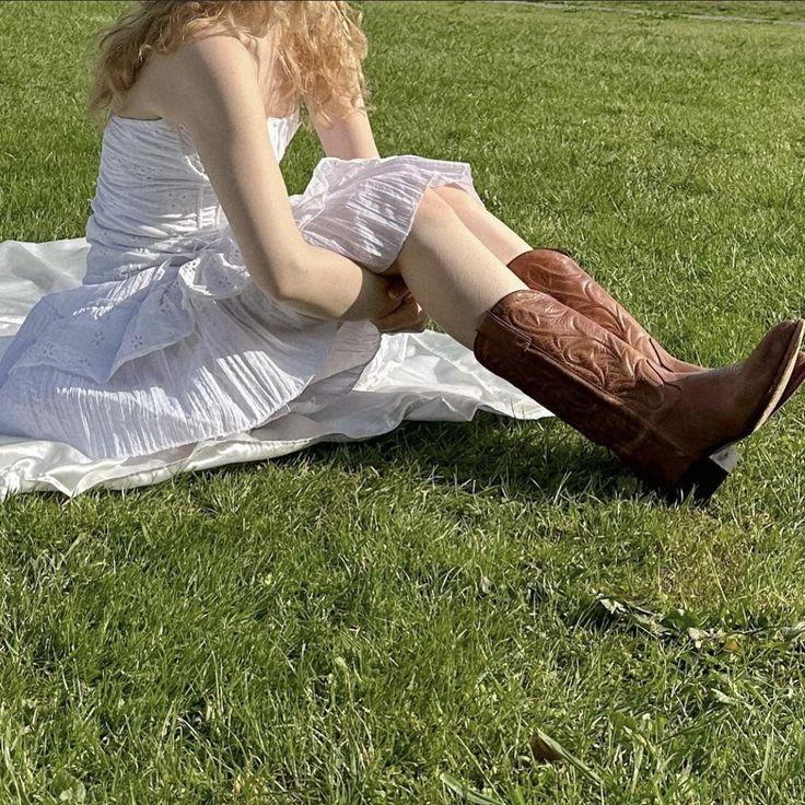 a woman in white dress sitting on the grass with her boots up to her knees