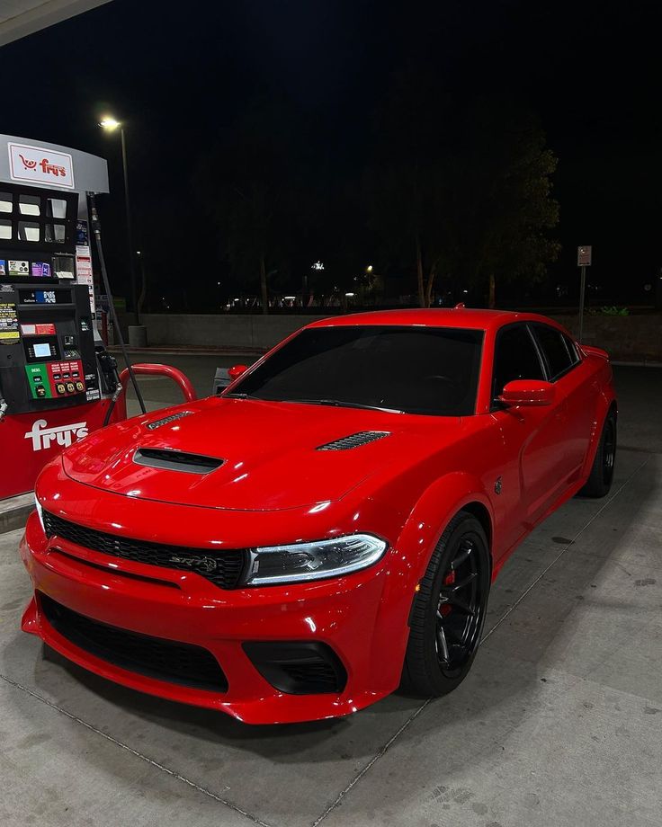 a red car is parked in front of a gas pump at night time, with the lights on