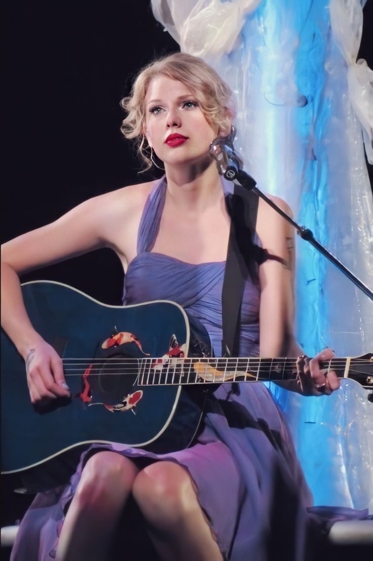 a woman in a purple dress is holding a guitar and singing into a microphone while sitting on a chair
