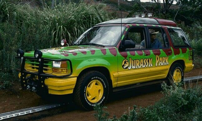 a green and yellow vehicle driving down a dirt road next to trees, bushes and plants