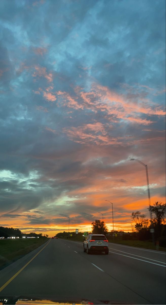 a car driving down the road at sunset with clouds in the sky and trees on either side