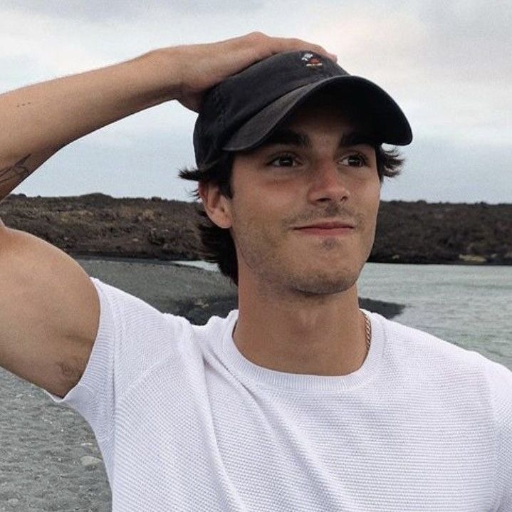 a man in a white shirt and black hat is standing by the water with his hands on his head