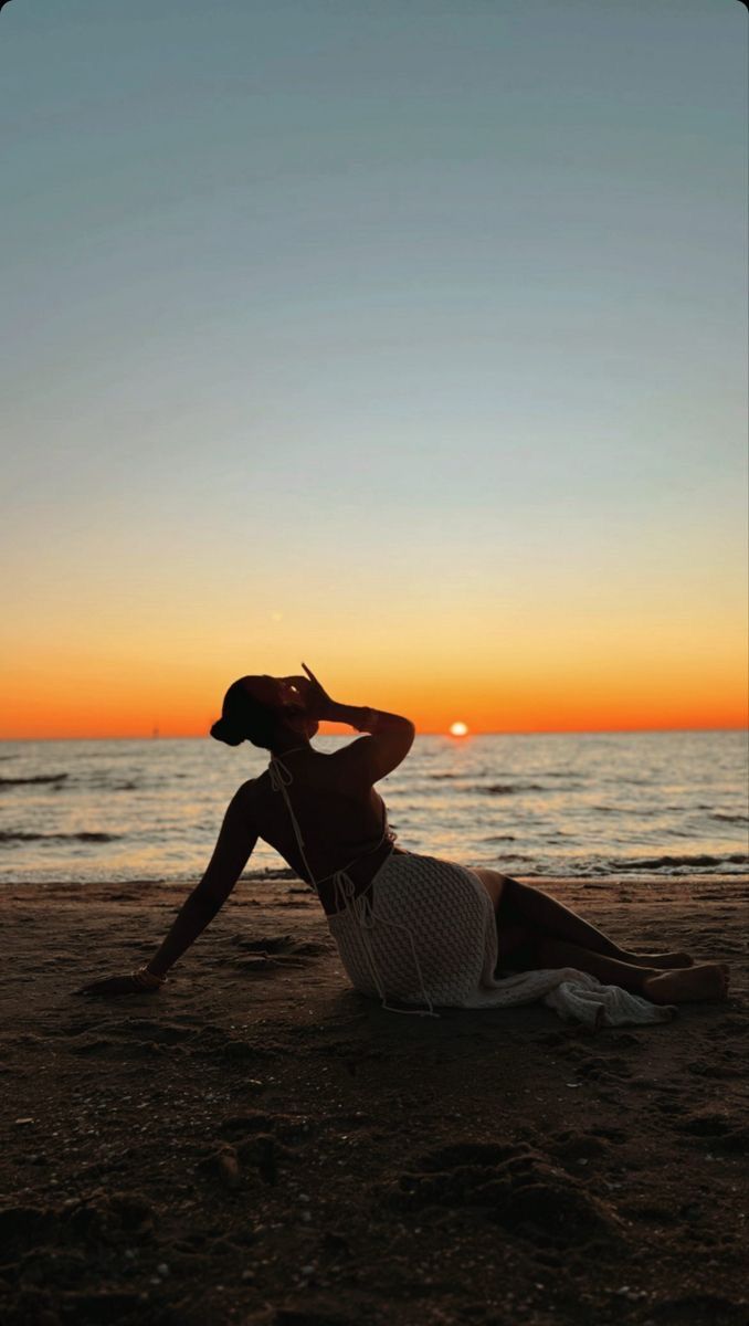 a woman sitting on top of a sandy beach next to the ocean at sun set