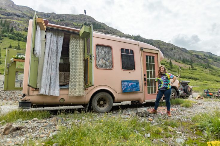 a woman standing in front of an old pink trailer with curtains on the door and windows