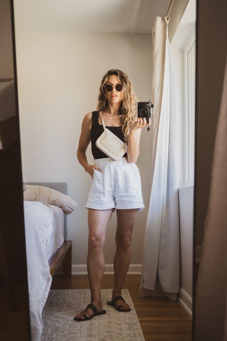 Ashley wearing a black tank, white linen shorts, black sandals, a beige belt bag, and tortoiseshell sunglasses Outfit With Linen Shorts, Olive Linen Shorts Outfit, Dinner Shorts Outfit, Tailored Black Shorts Outfit, White Shorts Black Top Outfit, Linen White Shorts, Summer Black And White Outfits, Linen White Shorts Outfit, Shorts And Tank Top Outfits Summer