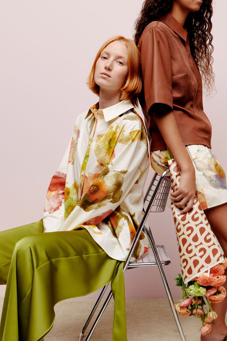 two women sitting next to each other in front of a pink wall and one is wearing a green skirt