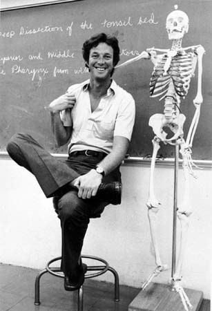 a black and white photo of a man sitting in front of a chalkboard with a skeleton on it