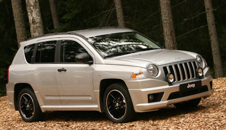 a silver jeep parked in front of some trees and leaves on the ground with it's hood up