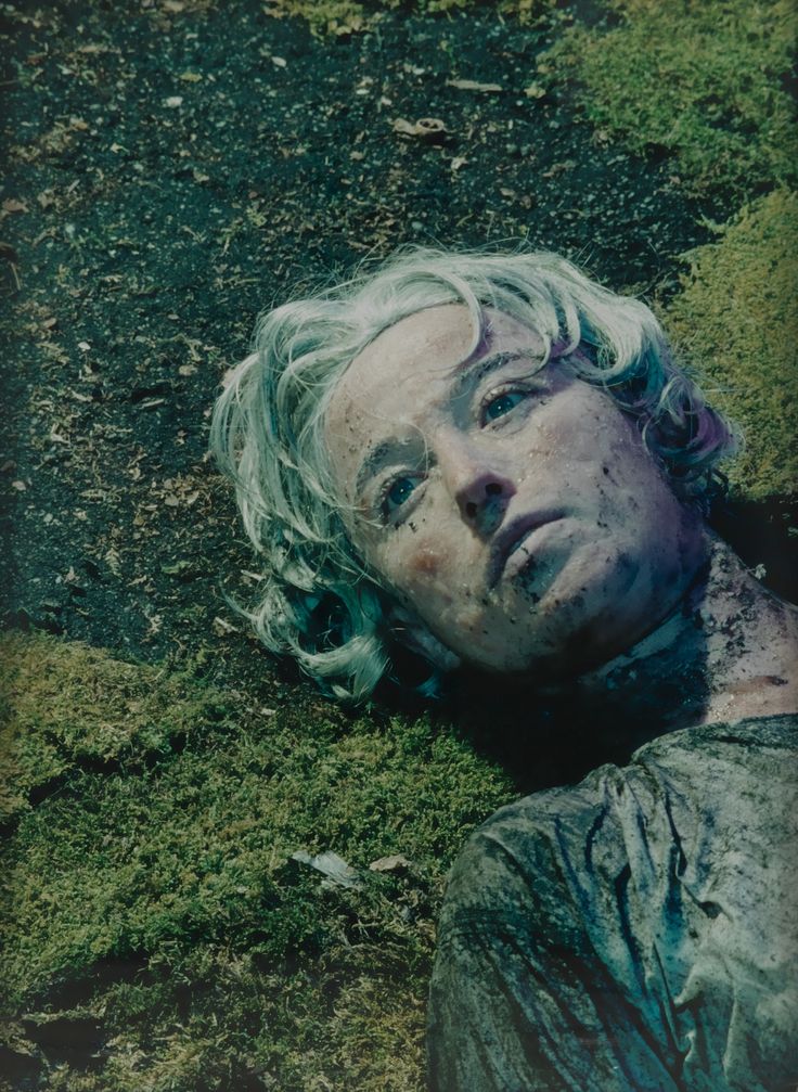 a man with white hair laying on the ground next to some green grass and dirt