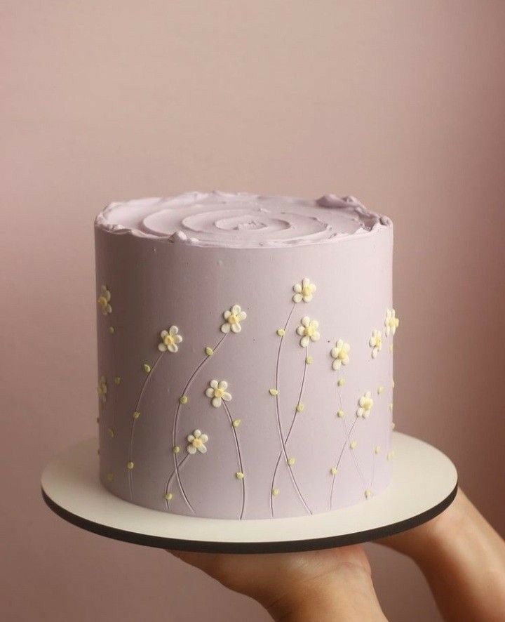 a hand holding a cake with white frosting and flowers on the top, in front of a pink background