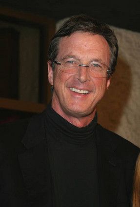 a man wearing glasses and a black turtle neck sweater smiles at the camera while standing next to a woman