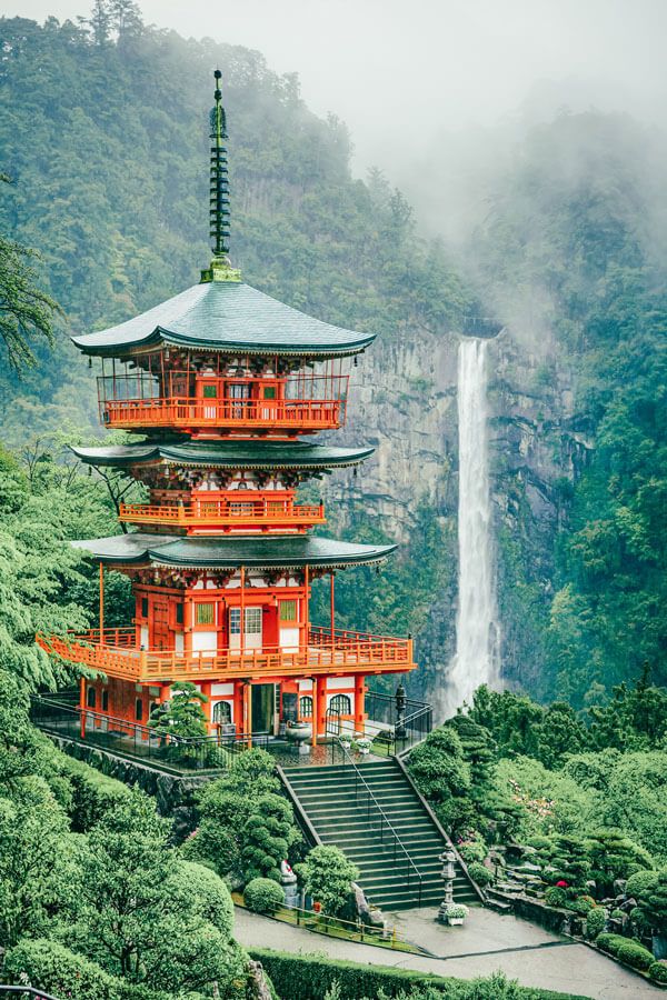 a tall red building sitting on top of a lush green hillside next to a waterfall