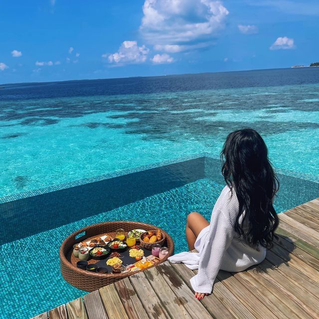 a woman sitting on a dock looking out at the ocean with food in front of her