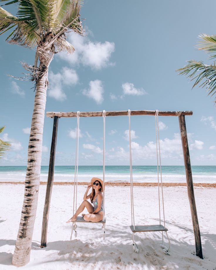 a woman sitting on top of a swing next to a palm tree at the beach