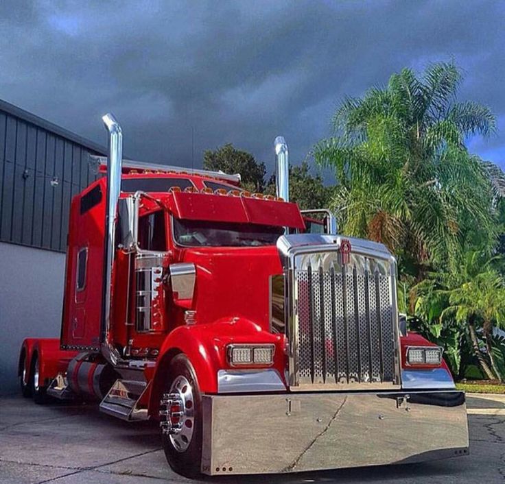 a red semi truck parked in front of a building with palm trees and dark clouds