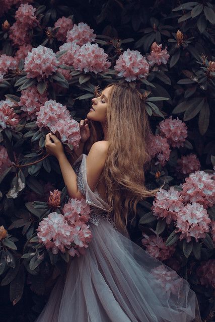 a woman with long hair standing in front of flowers wearing a gray dress and holding a pink flower