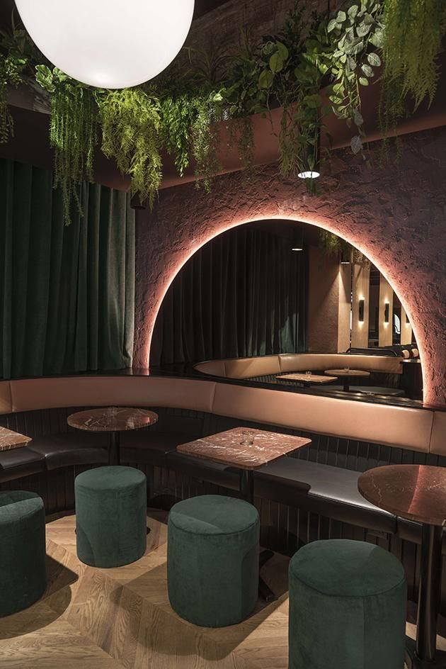 the interior of a restaurant with round tables and green velvet stools, plants hanging from the ceiling