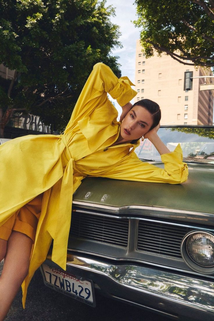 a woman leaning on the hood of an old car wearing a yellow dress and heels