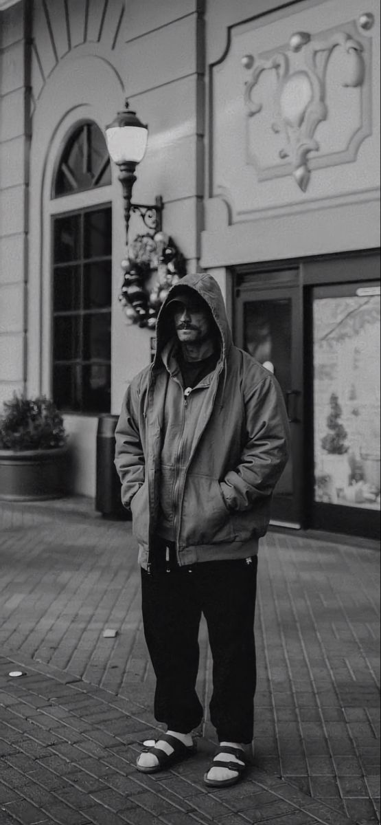 black and white photograph of man in hoodie standing on the sidewalk with his hands in his pockets