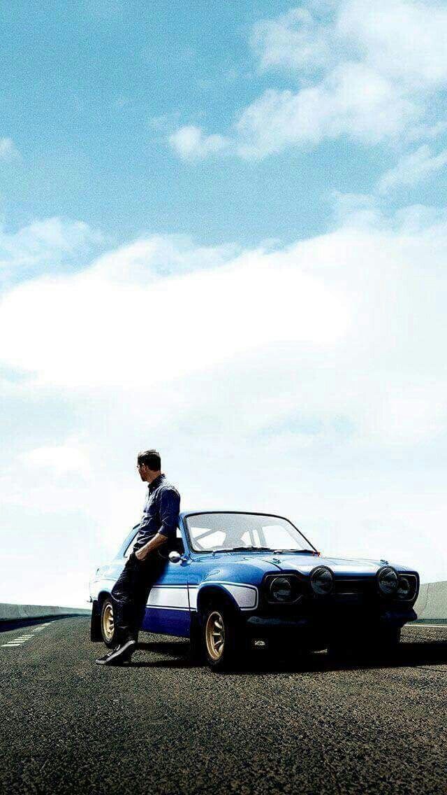 a man sitting on the hood of a blue car in front of a sky with clouds