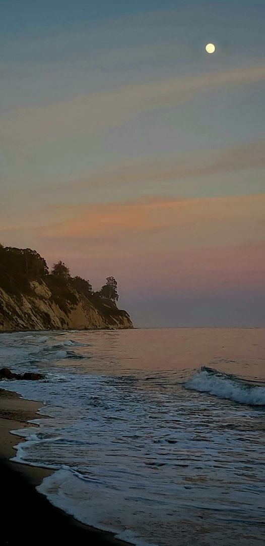 the moon is setting over the ocean with waves crashing on the shore and trees in the distance