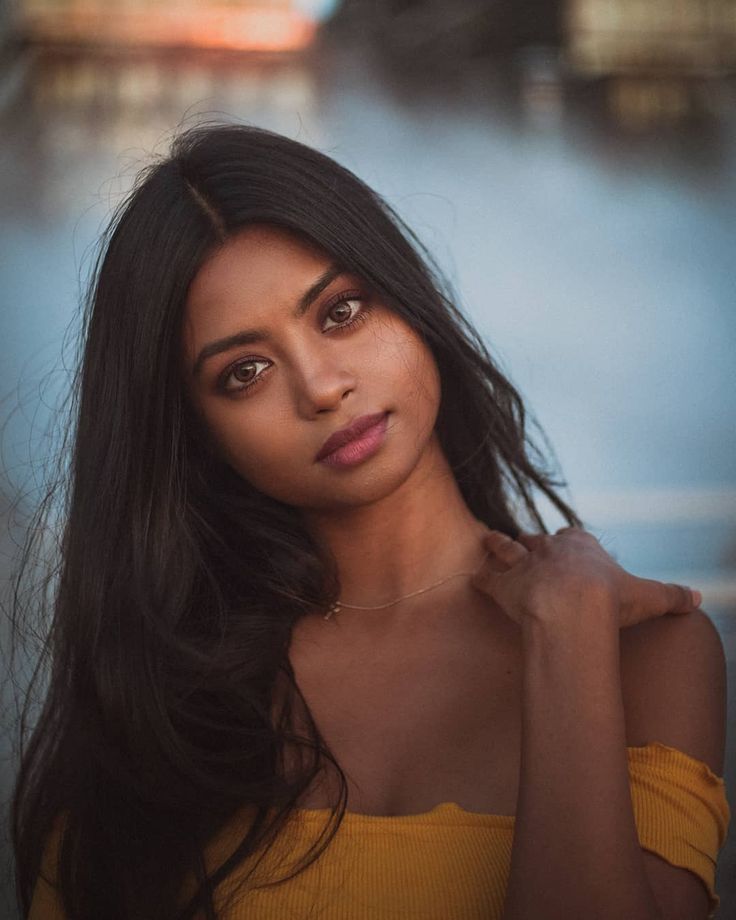 a woman with long dark hair and brown eyes poses for a photo in front of the water