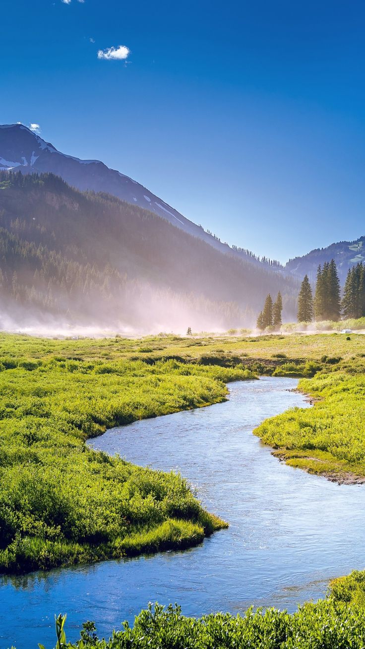 a river running through a lush green field next to a mountain covered in fog and mist