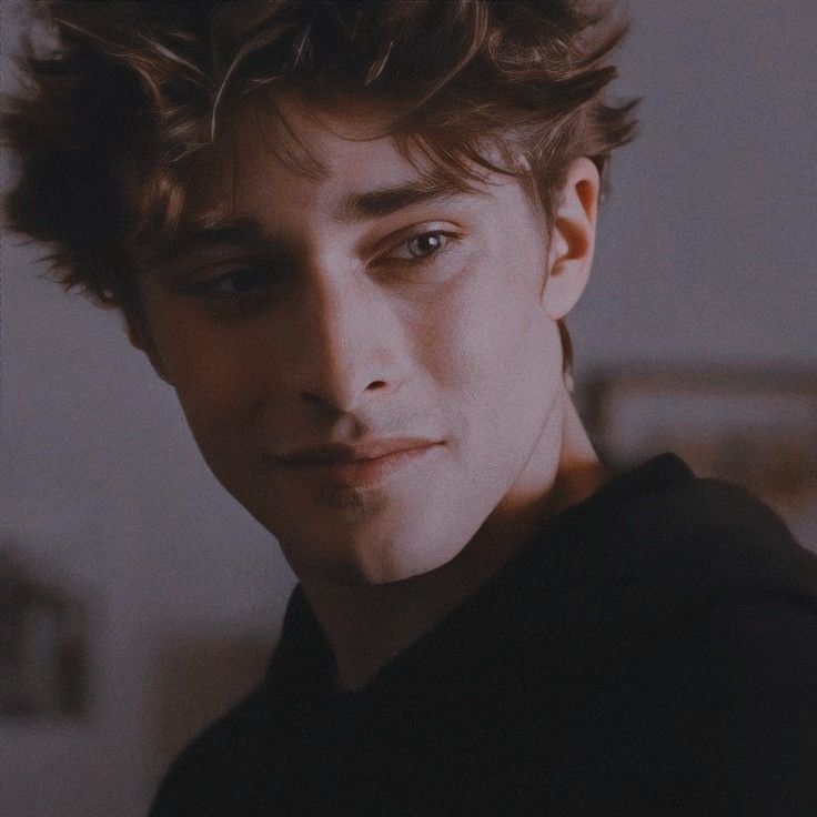 a young man with curly hair and blue eyes looks into the camera while wearing a black shirt