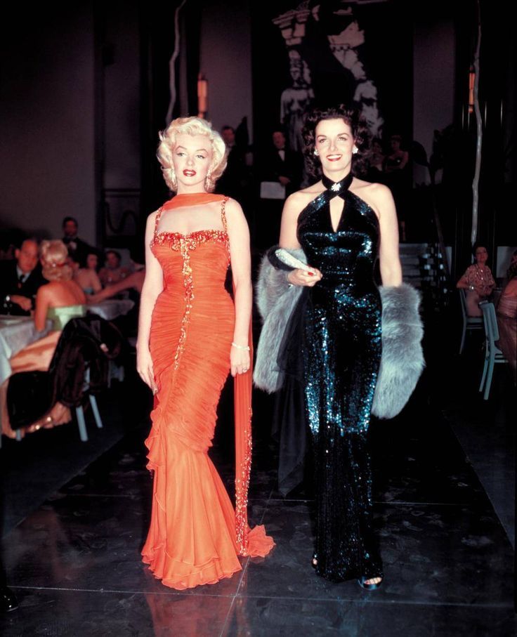 two women in evening dresses standing next to each other at a formal event, one wearing an orange dress and the other black