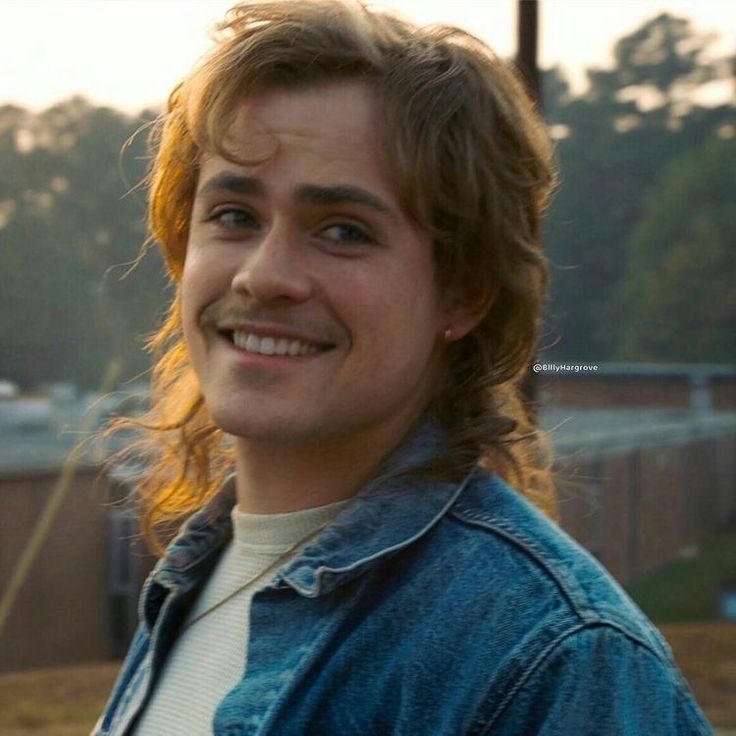 a young man with long hair smiles at the camera while wearing a denim jacket and white shirt