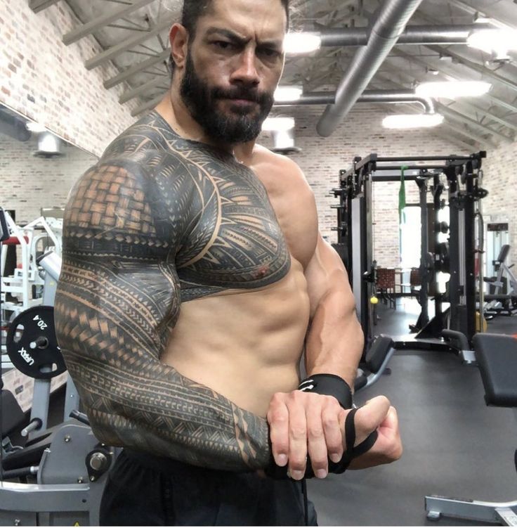 a man with tattoos on his arms and chest standing in a gym holding a barbell