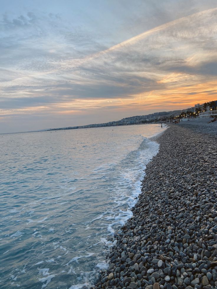 the beach is lined with rocks and pebbles as the sun sets over the water in the distance