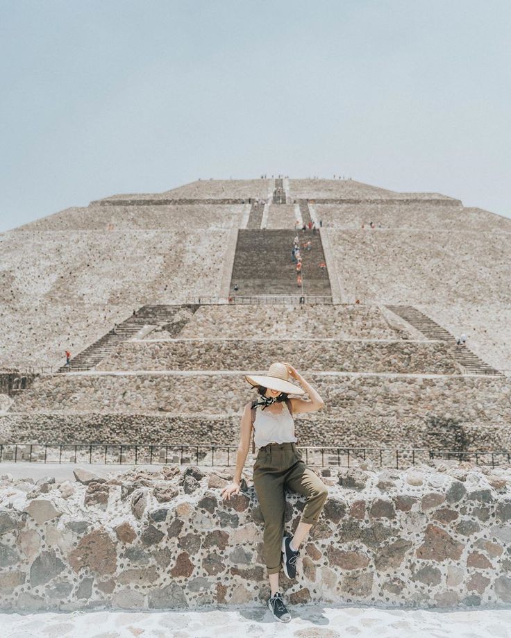 a woman standing in front of the pyramid of the sun wearing a straw hat and green pants