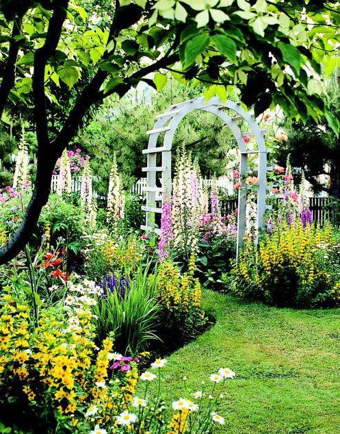 a garden with lots of flowers and trees in the background, including an arch shaped trellis