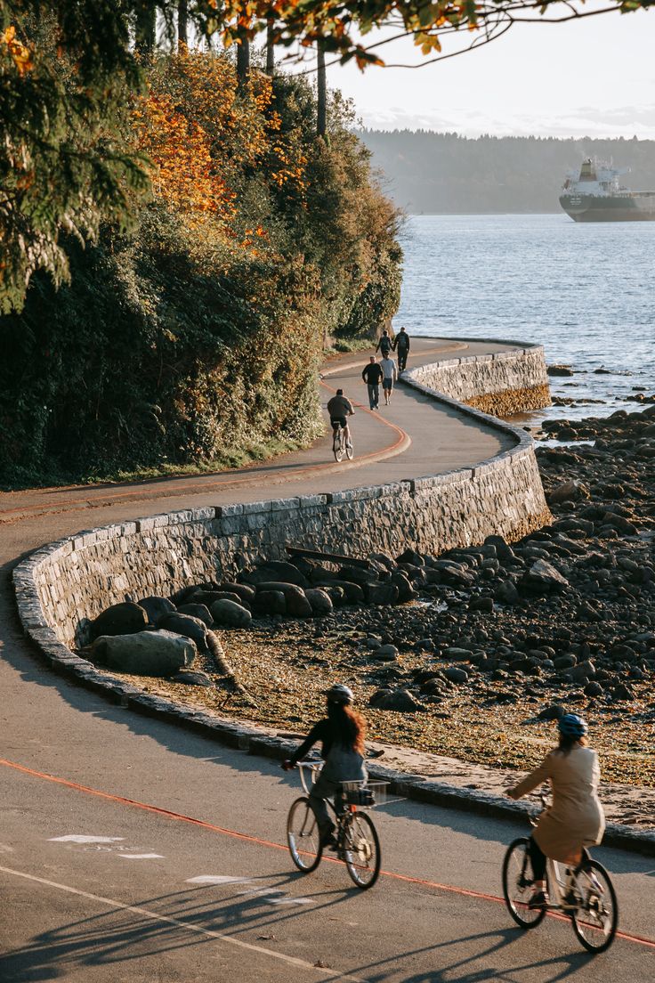 two people riding bikes on a paved road near the water