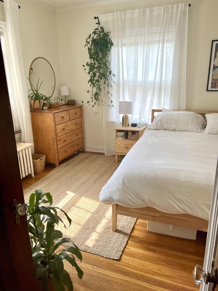 a bed sitting in a bedroom next to a window with white curtains and a potted plant on top of it