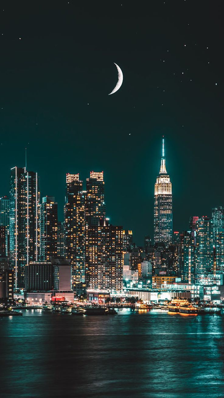 the city skyline is lit up at night, as seen from across the water with moon and stars in the sky