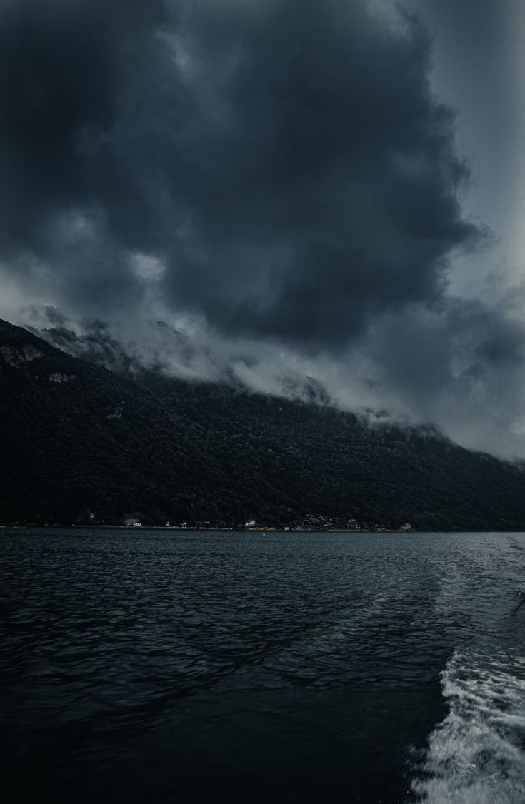 a boat traveling on the water under a cloudy sky with mountains in the background and dark clouds above