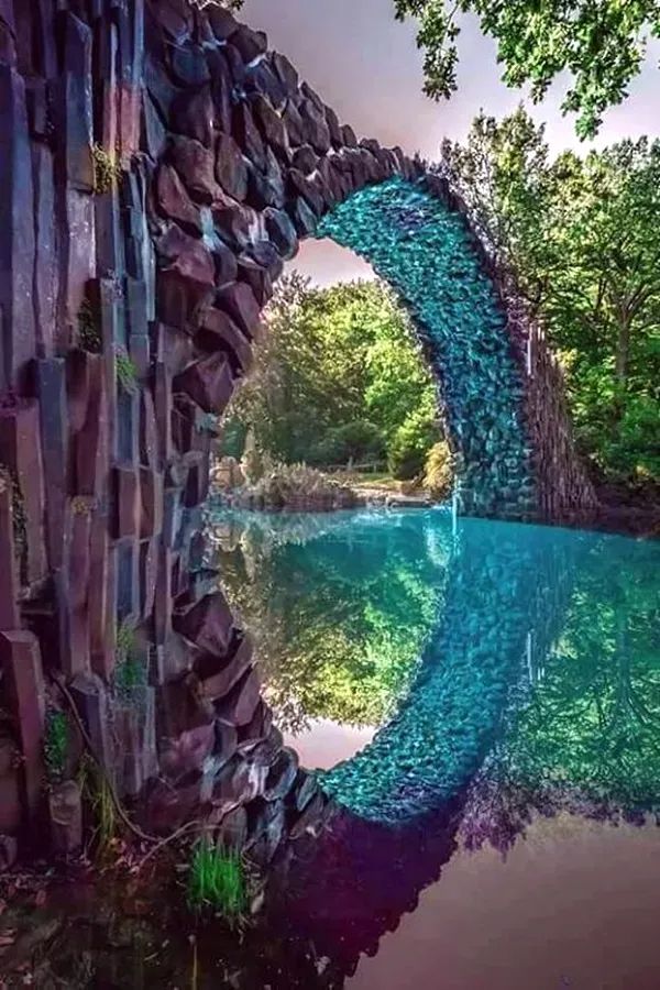 a stone bridge over a river surrounded by trees and rocks, with the reflection of it in the water
