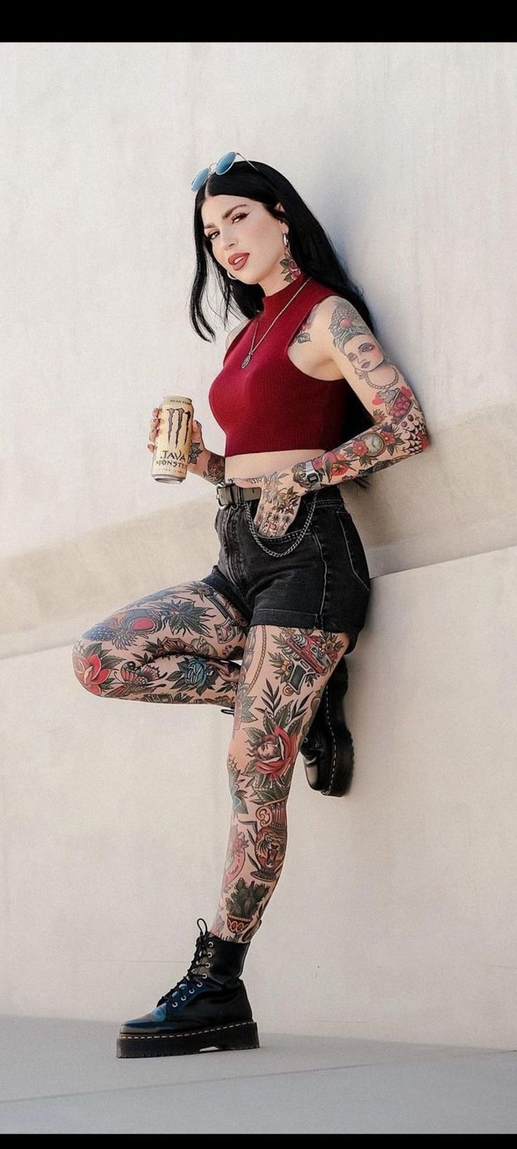 a woman with tattoos is leaning against a wall and holding a cup in her hand