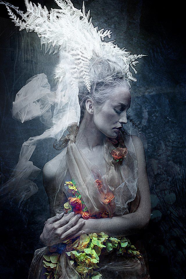 a woman with feathers on her head is sitting in front of a dark background and holds flowers