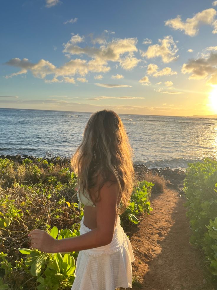 a woman walking down a dirt path next to the ocean at sunset or sunrise with her hair blowing in the wind