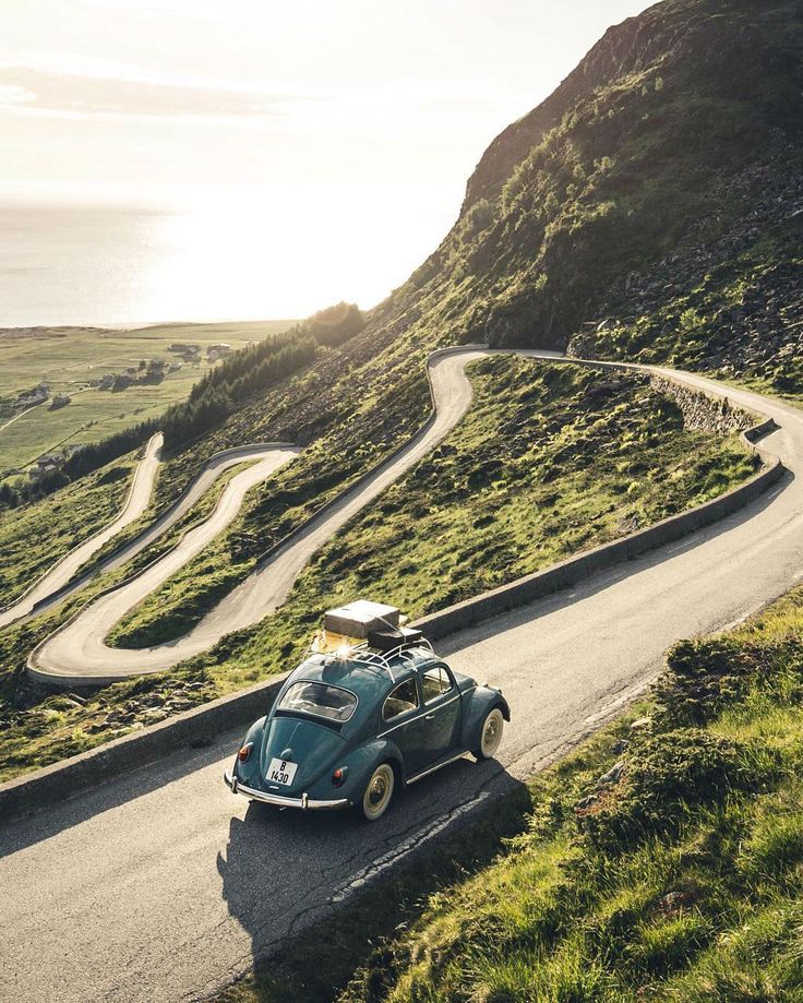 an old blue car driving down a winding road next to the ocean and grassy hills