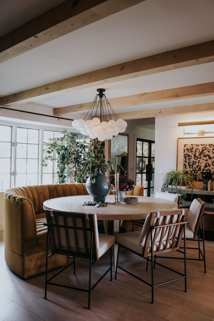 a dining room table with chairs and a couch in front of the window is surrounded by potted plants